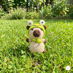 A crocheted plush toy dog wearing a green frog hat and scarf, standing in a grassy garden with blurred flowers in the background.
