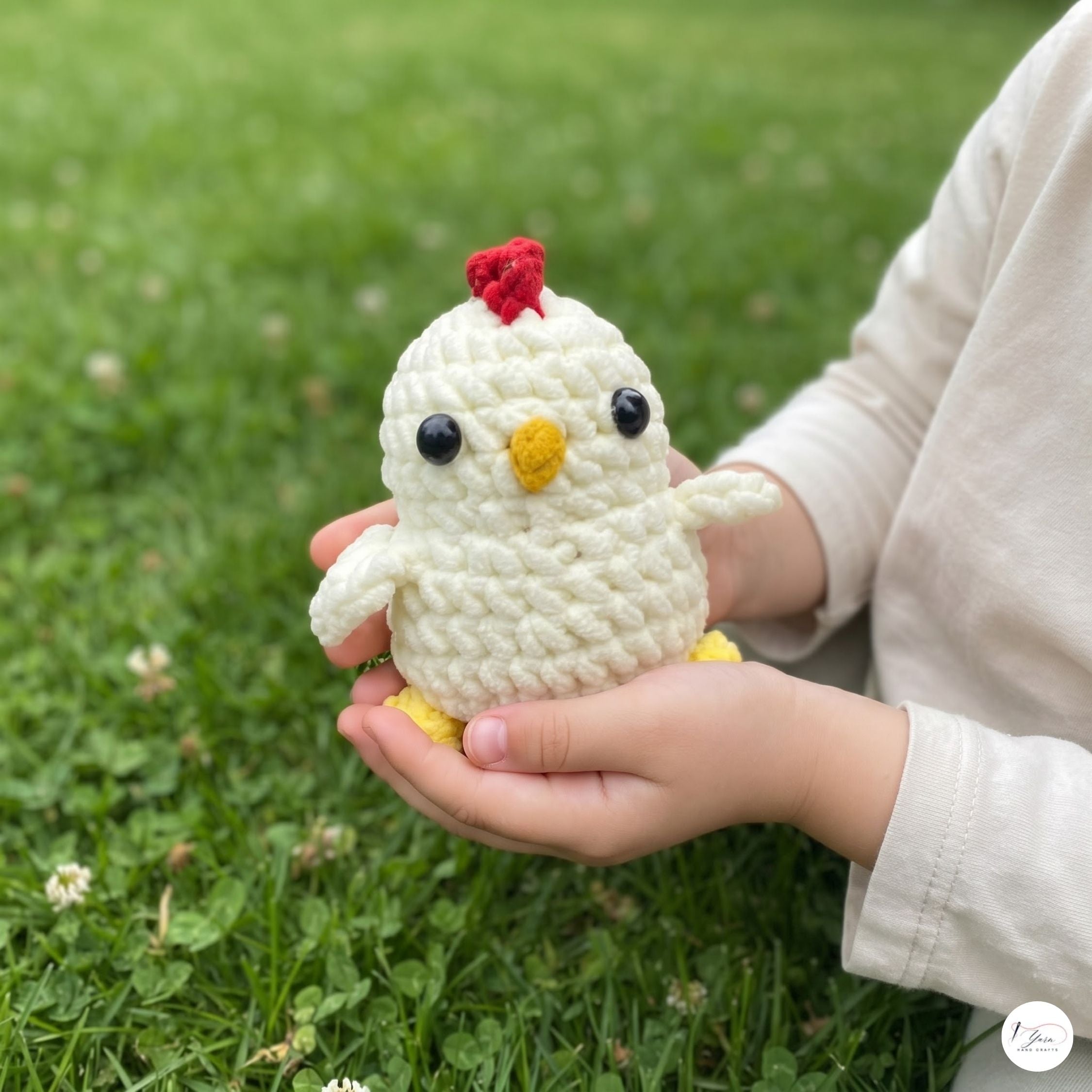 A small crocheted white chicken toy with a red comb, yellow beak and feet, and black bead eyes is being held in a person's hands outdoors on a green grass background.