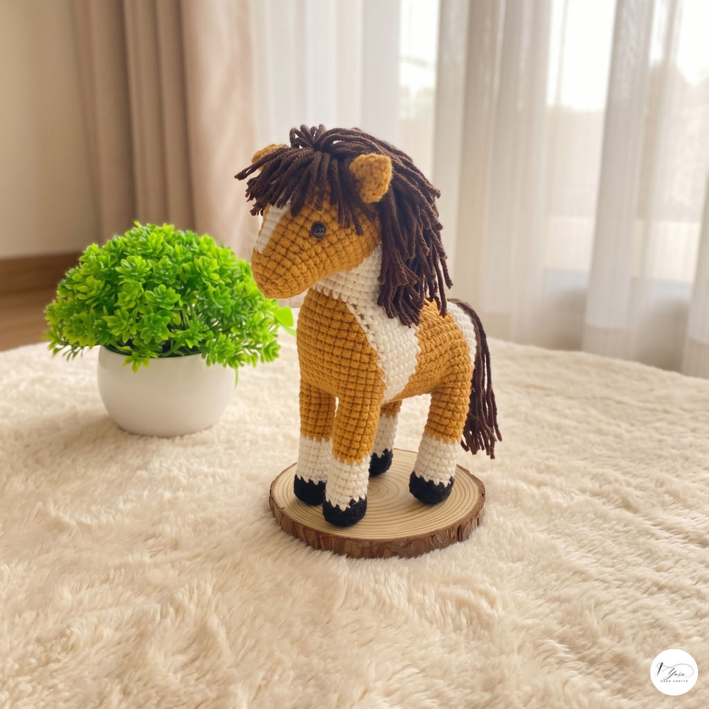 A crocheted amigurumi horse toy standing on a round wood slice base, placed on a cream shag rug beside a small potted green plant in a softly lit indoor setting.