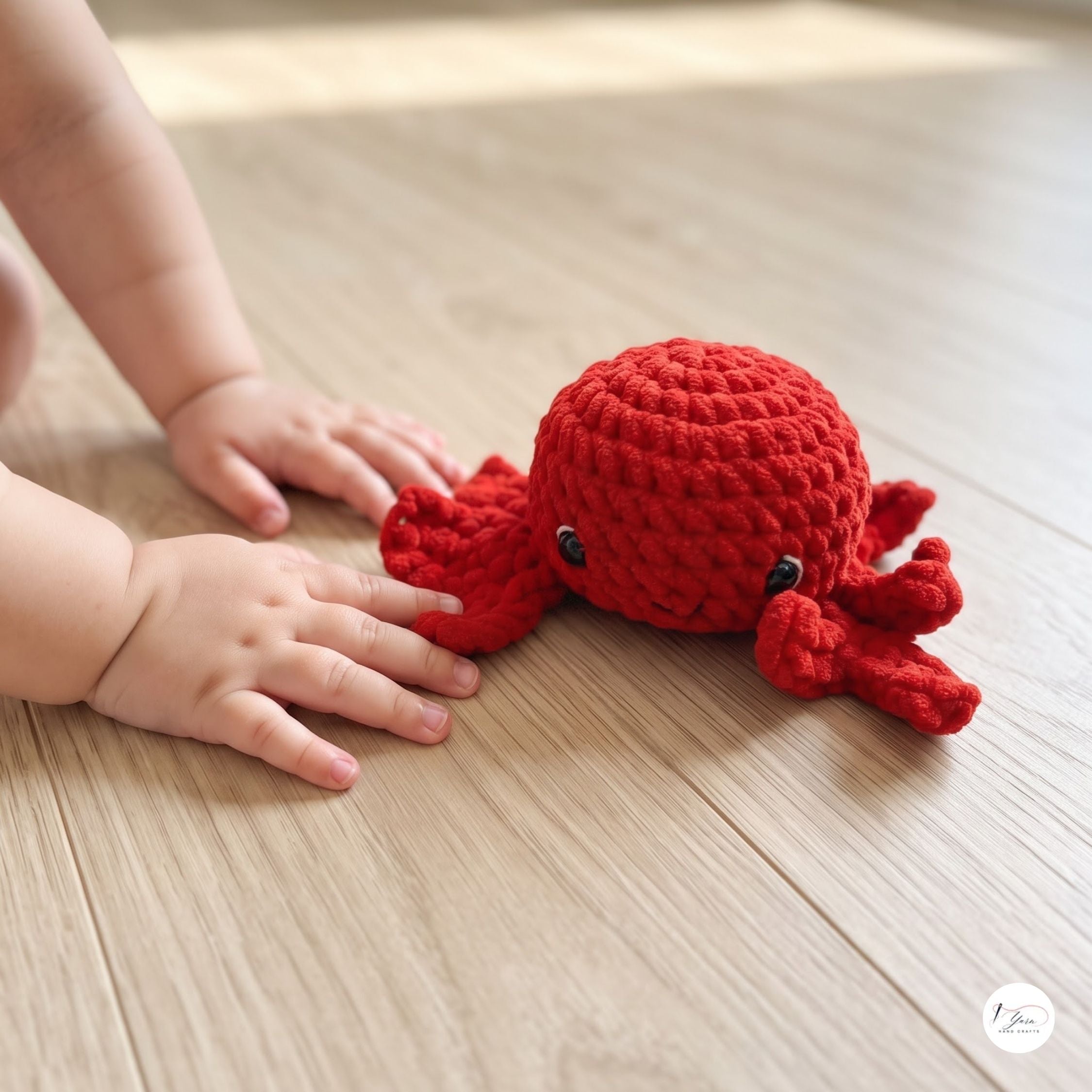 A baby's hands and arms are reaching toward a small red crocheted octopus toy on a light wood floor.