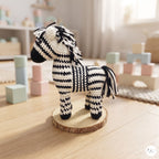 A crocheted zebra toy doll with a black and white striped pattern, standing on a round wooden slice coaster in a softly blurred children's playroom setting.