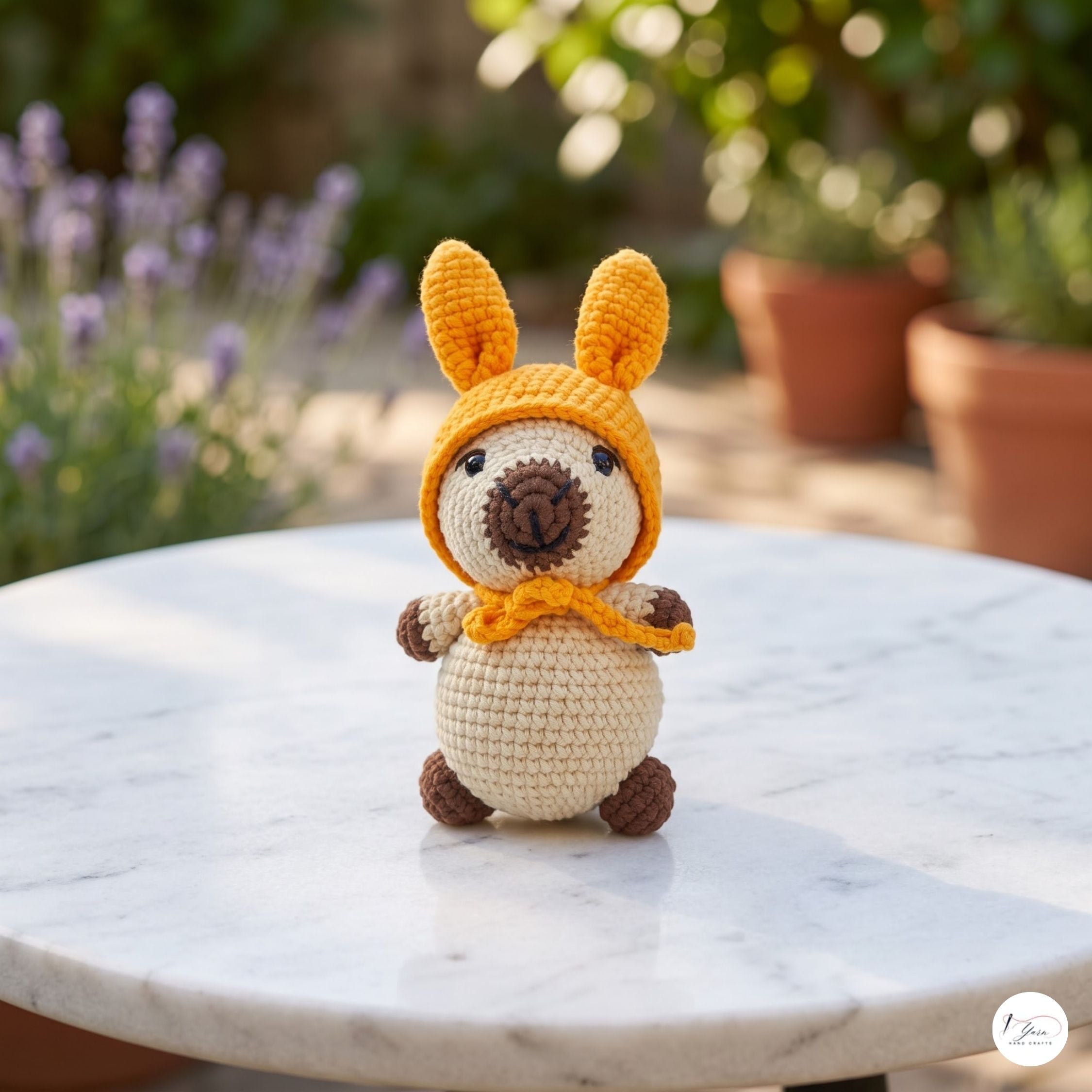A crocheted plush toy kangaroo wearing a yellow hood and matching scarf, sitting on a white marble table with a blurred garden background of potted plants and lavender.