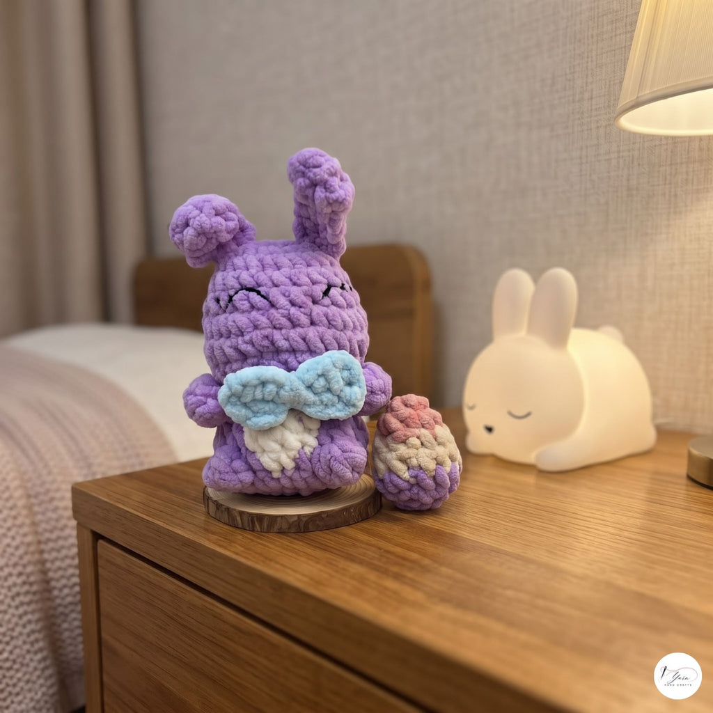 A crocheted purple bunny figurine holding a blue bow and standing beside a small pink-and-white pouch, placed on a wooden bedside table in a softly lit bedroom. A white bunny lamp sits in the background.