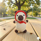 A crocheted teddy bear toy wearing a red hat and red scarf, standing on a wooden outdoor table with a blurred park background.