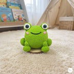 A green crocheted frog toy with large white eyes and a black stitched smile, sitting on a small round wooden base on a cream-colored carpet in a child's room.