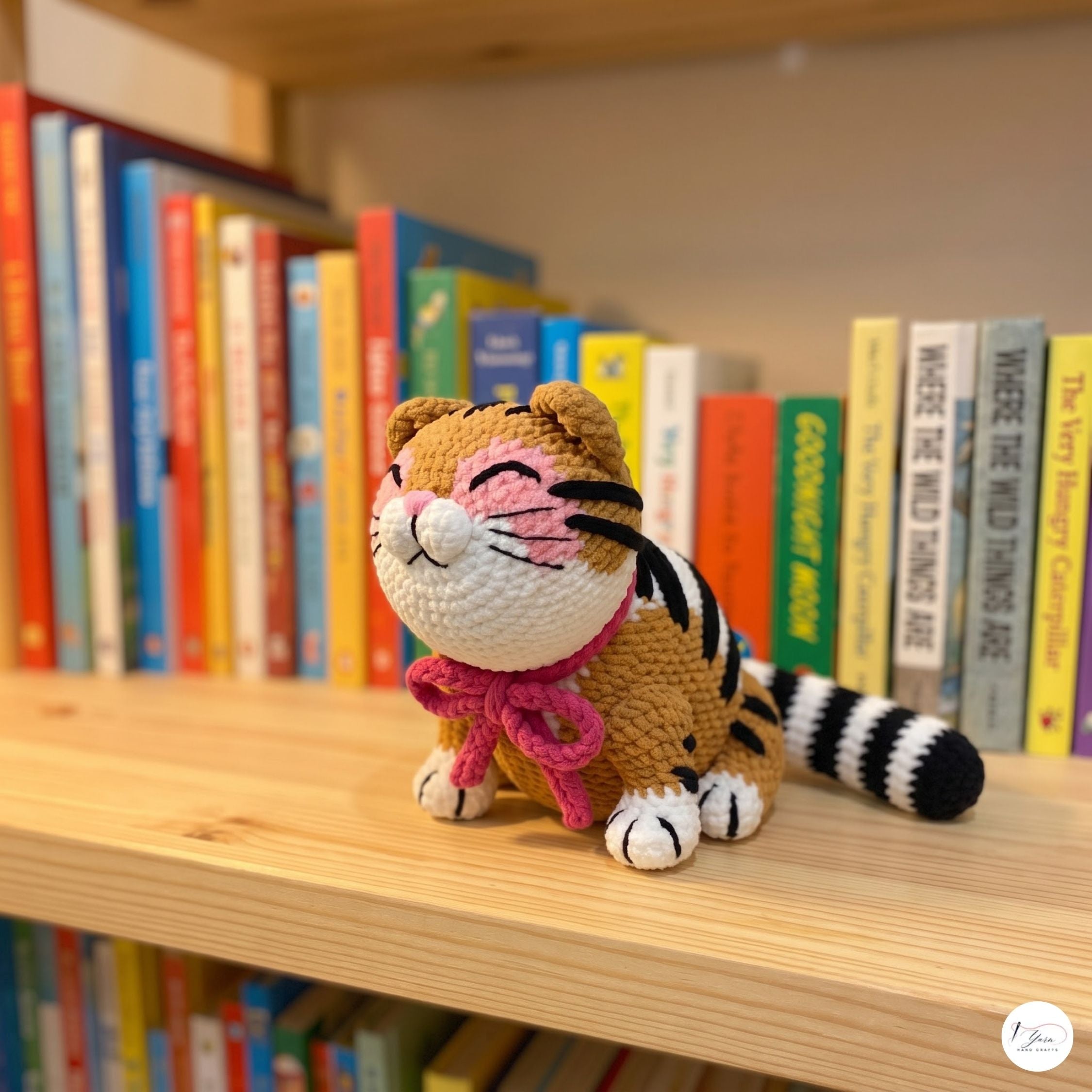A crocheted tiger plush toy with orange and black stripes, a pink bow, and a white face sits on a wooden bookshelf in front of colorful books.