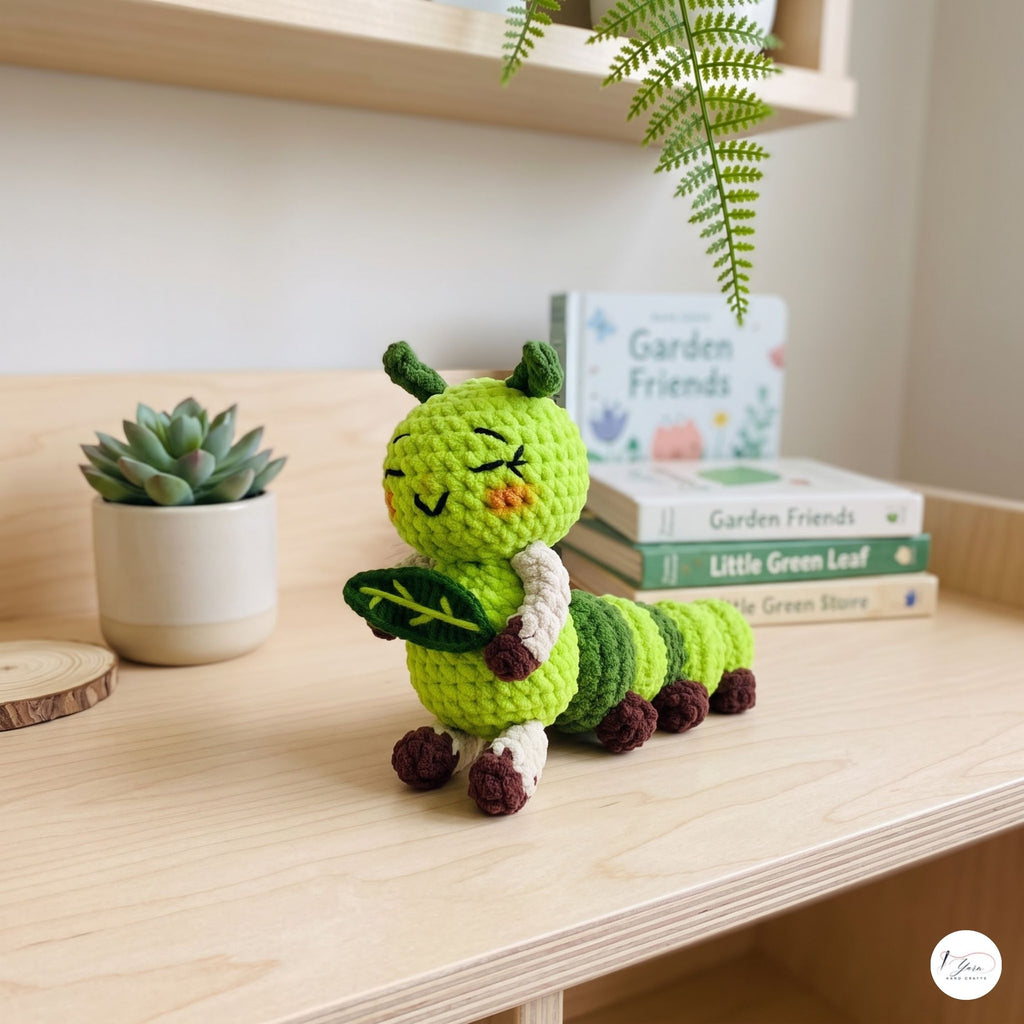 A crocheted caterpillar plush toy in bright green and yellow-green, sitting on a light wood shelf next to a small potted succulent and a stack of books.