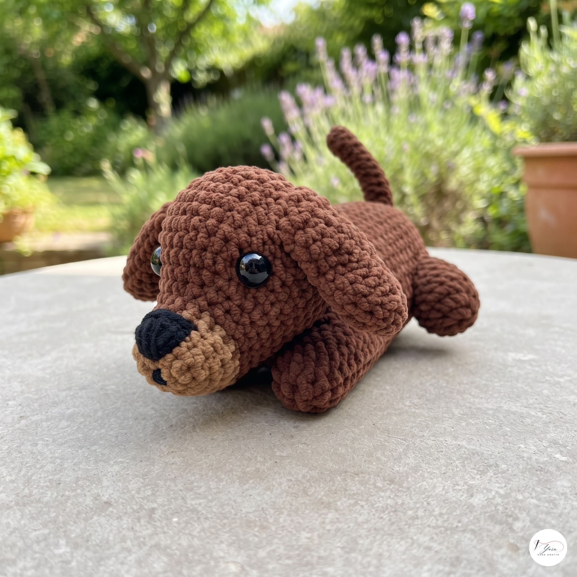 A crocheted brown puppy toy with a tan snout and black nose, lying on a light gray stone surface with a blurred garden background.