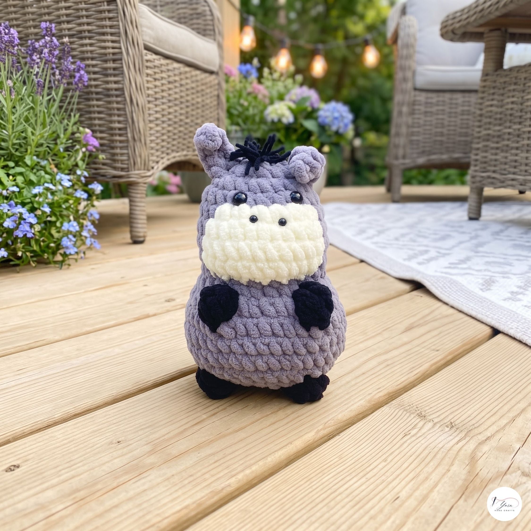 A small crocheted hippo plush toy sits on a wooden deck outdoors, with wicker patio furniture and flowers blurred in the background.