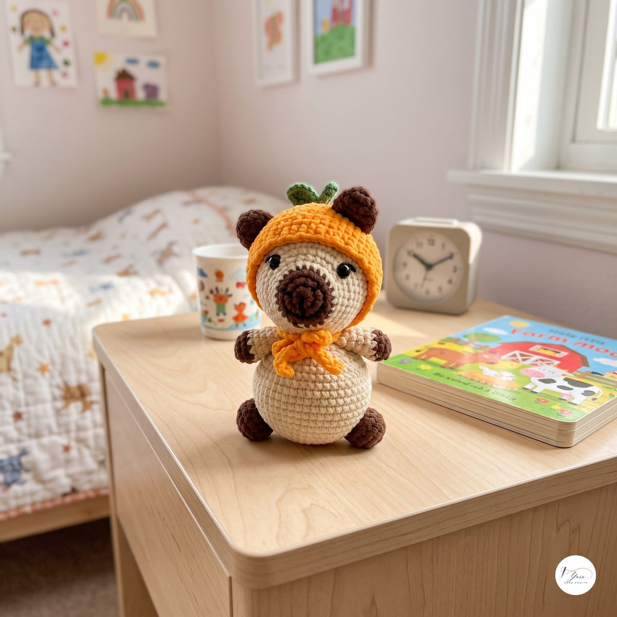 A crocheted plush toy with a cream body, brown ears and feet, and an orange pumpkin hat, styled on a light wood bedside table in a child's bedroom next to a mug, clock, and book.