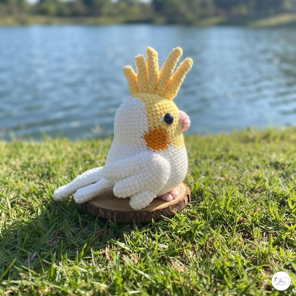 A crocheted amigurumi bird toy with a yellow crest, white body, and orange chest patch, sitting on a small wooden slice outdoors on grass with a lake in the background.