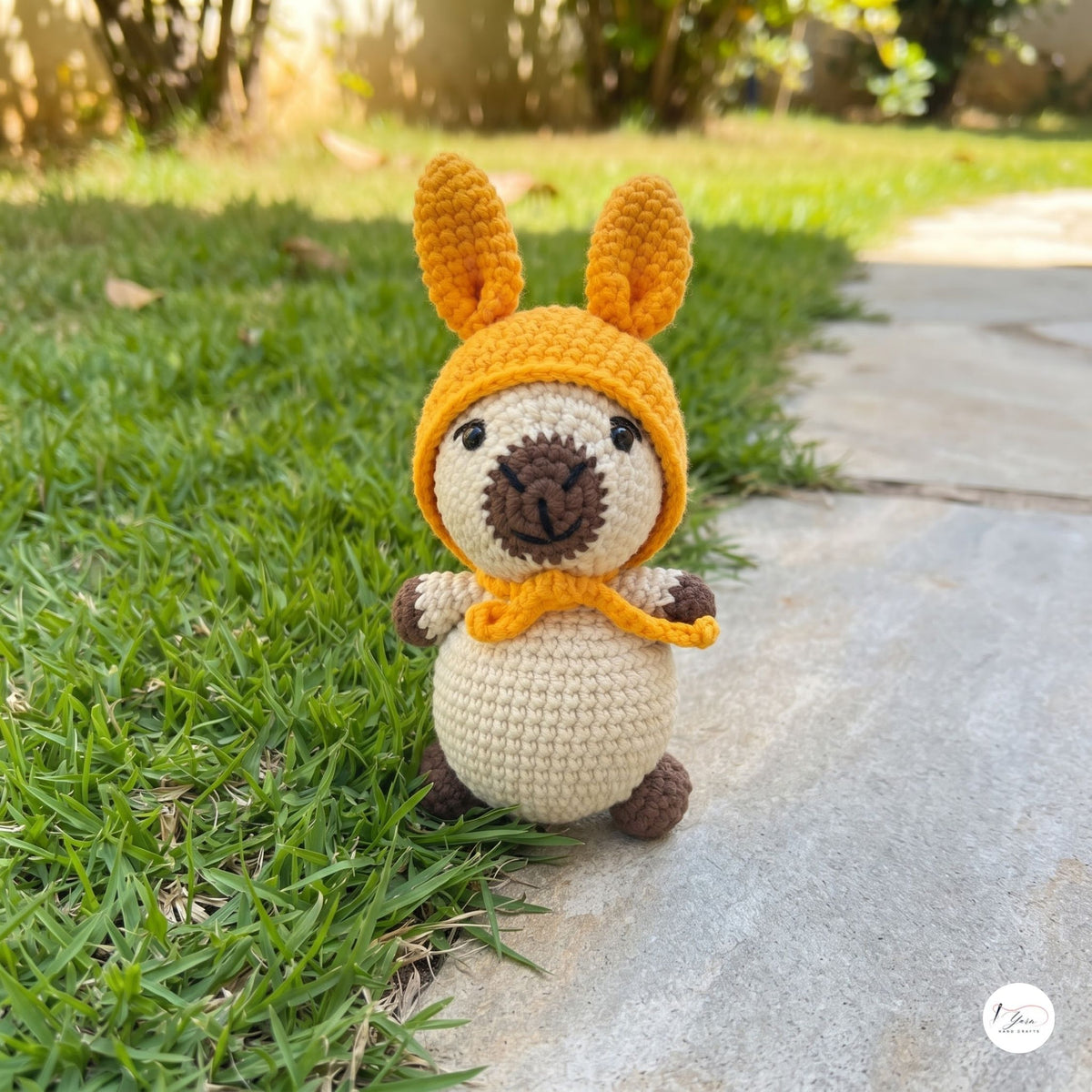 A small crocheted plush toy with long orange ears, a cream body, and brown accents sits on a stone path beside green grass outdoors.