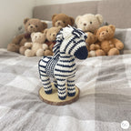 A crocheted zebra toy standing on a round wood slice coaster on a gray and white checkered bed, with a collection of teddy bears in the background.