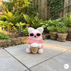 A cute handmade white crochet cat plush sitting on a stone patio in an outdoor garden setting, wearing a pink sweater, dark pink-framed sunglasses over its eyes, and holding a crocheted takeaway coffee cup.