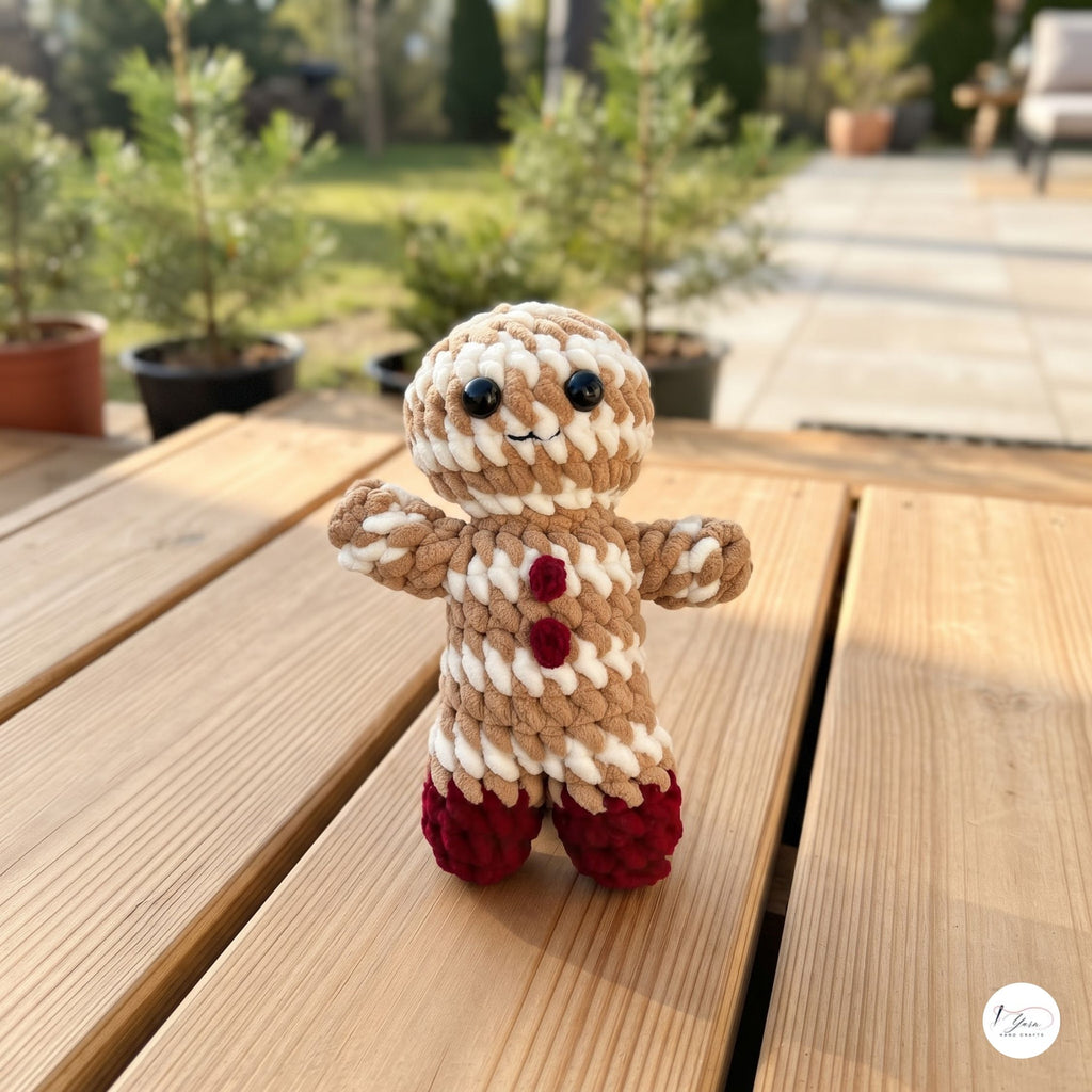 A small crocheted gingerbread person doll stands on a wooden table outdoors, with blurred potted plants and a patio walkway in the background.