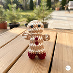 A small crocheted gingerbread person doll stands on a wooden table outdoors, with blurred potted plants and a patio walkway in the background.