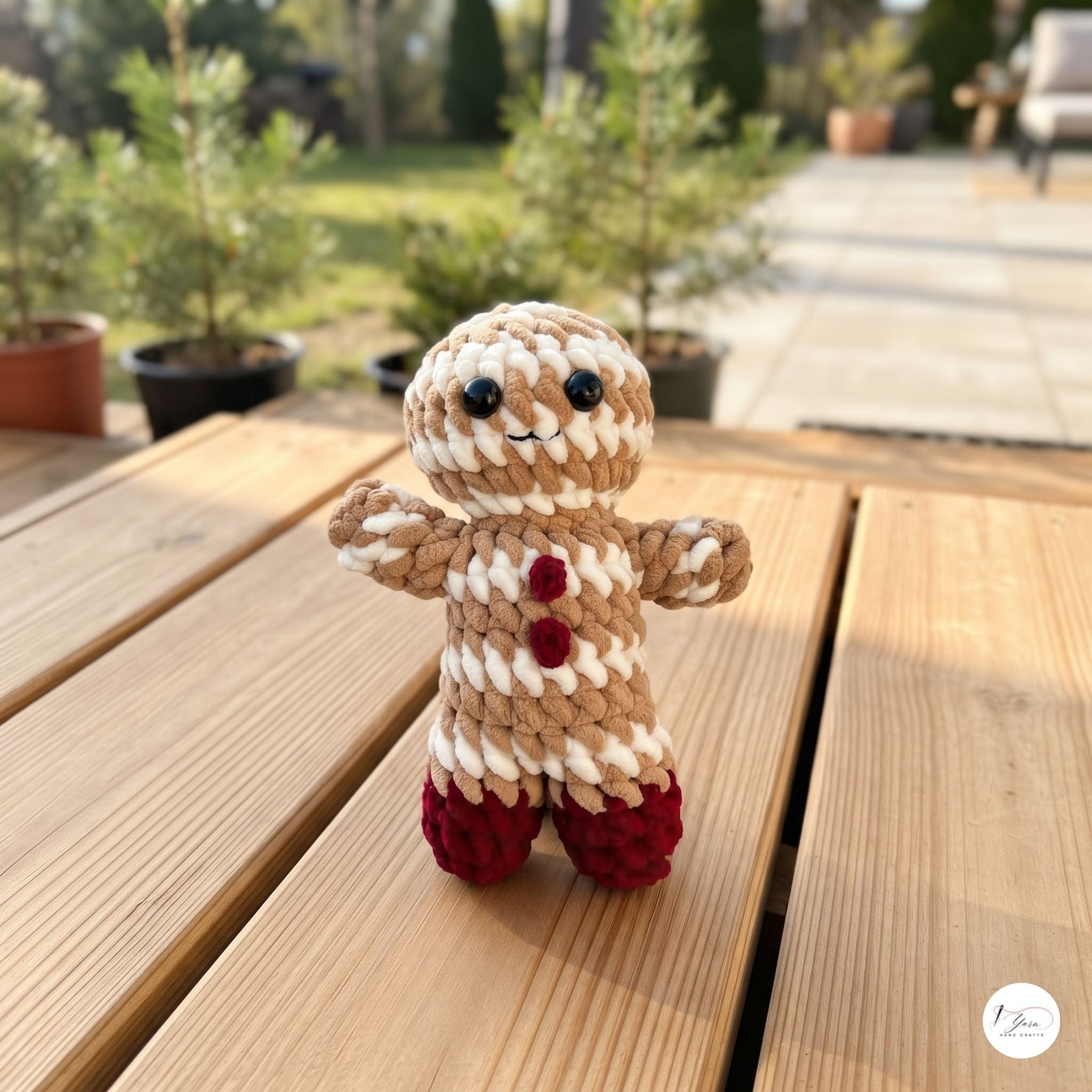 A small crocheted gingerbread person doll stands on a wooden table outdoors, with blurred potted plants and a patio walkway in the background.