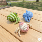 A plush handmade set of three crochet spider toys in mottled green and white, light blue, and pink and cream, arranged together on a sunlit wooden picnic table outdoors.