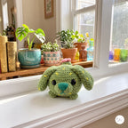A small crocheted plush bunny in light green sits on a white windowsill, with potted plants and books visible in the softly lit room behind it.