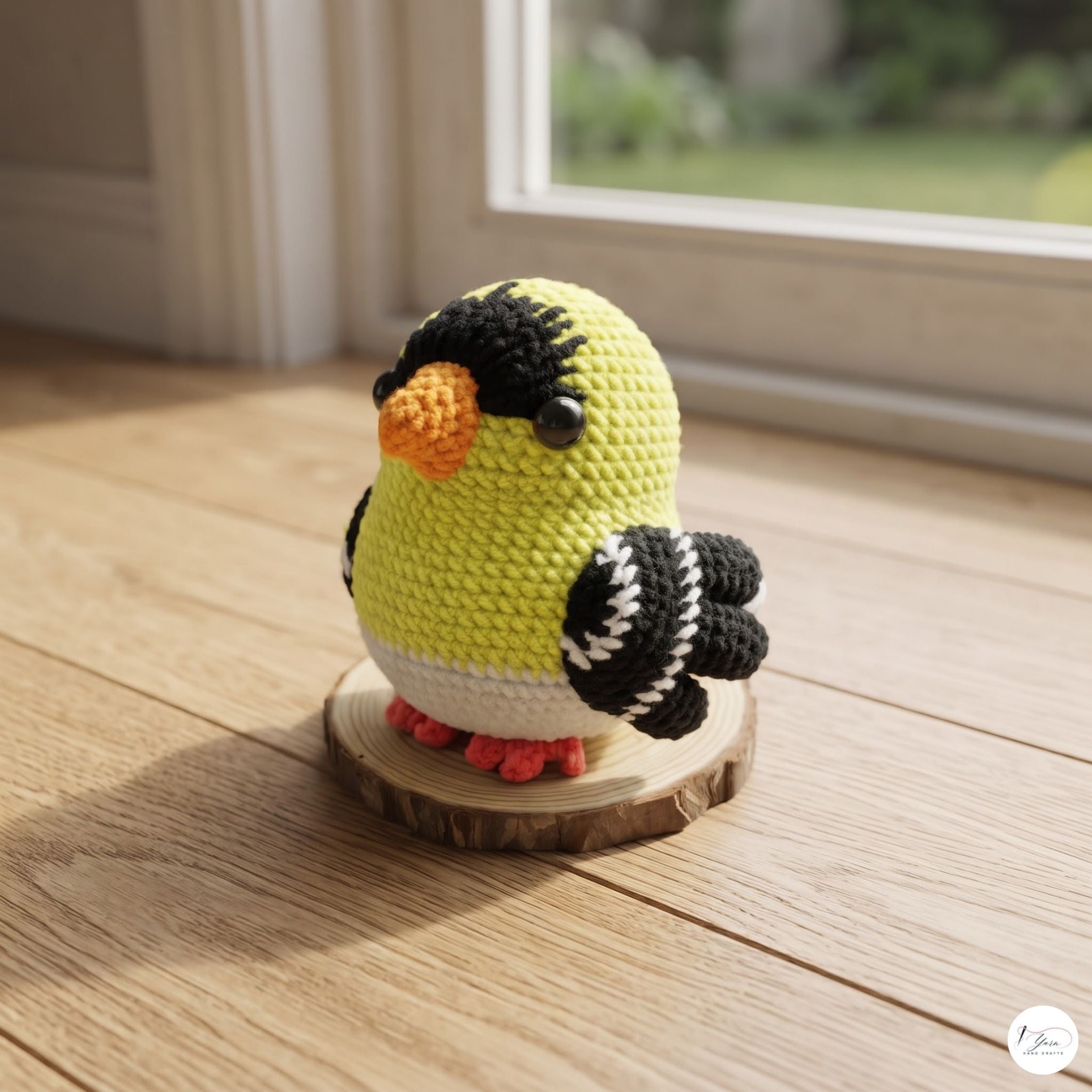 A crocheted bird figurine with a yellow body, black and white wings, and an orange beak, standing on a small round wooden slice on a light wood floor near a window.