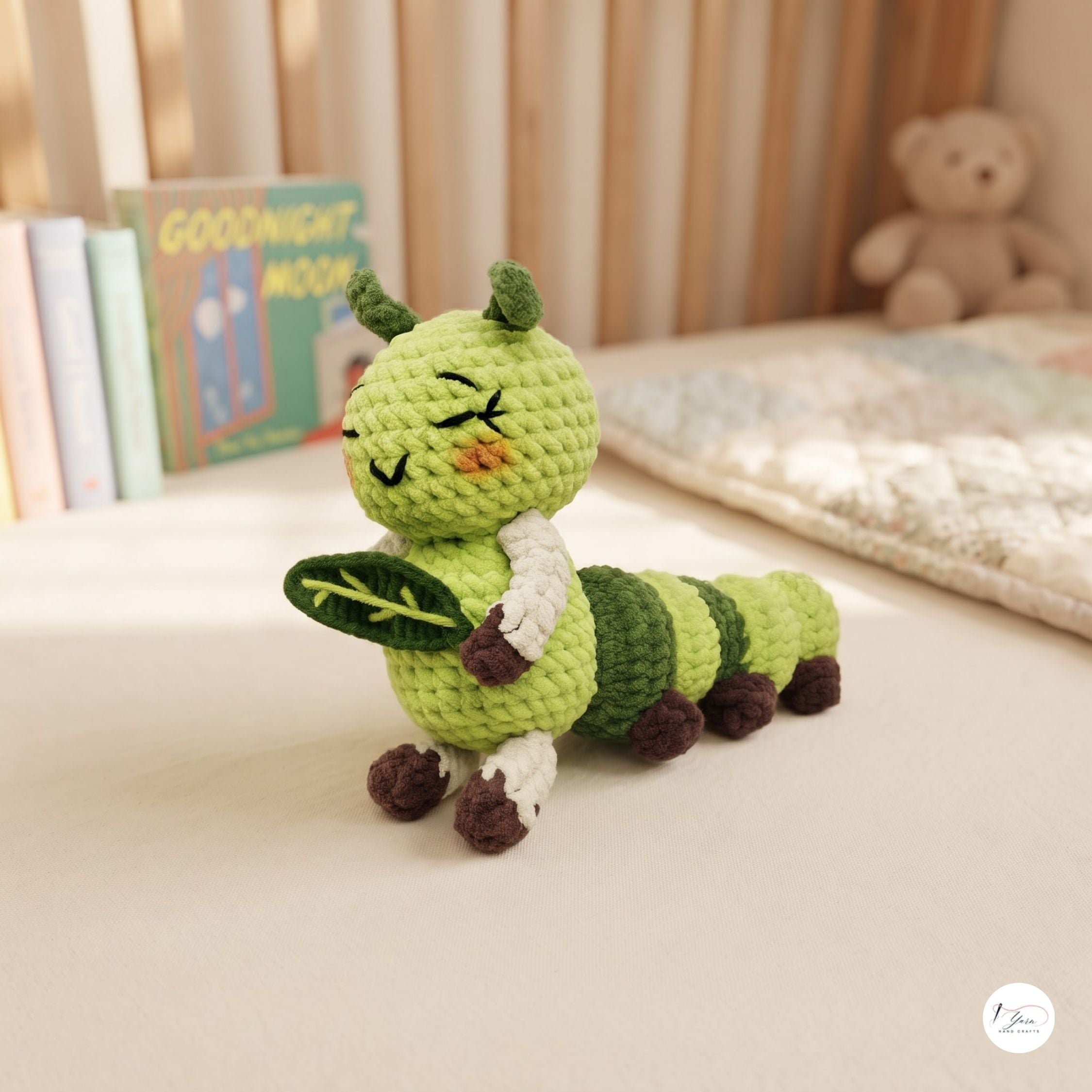 A handmade crocheted caterpillar toy in shades of green, brown, and white, sitting on a light beige surface with a softly blurred nursery background containing books and a teddy bear.