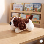 A crocheted plush puppy toy with brown and white yarn sits on a light wood table in front of a wooden bookshelf filled with children's books.