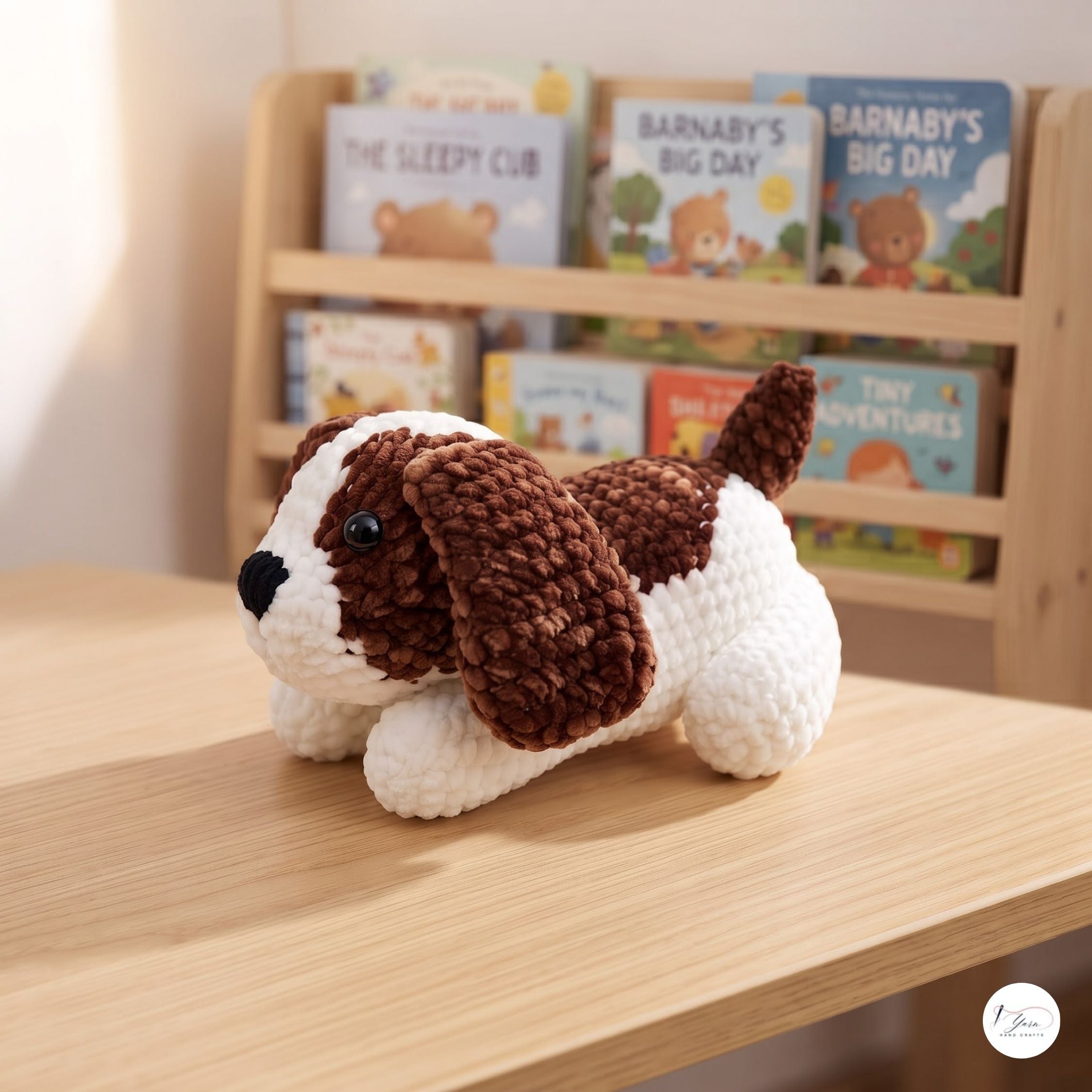A crocheted plush puppy toy with brown and white yarn sits on a light wood table in front of a wooden bookshelf filled with children's books.