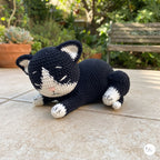 A crocheted black and white cat toy lying down on a beige stone patio with a garden bench and plants in the background.
