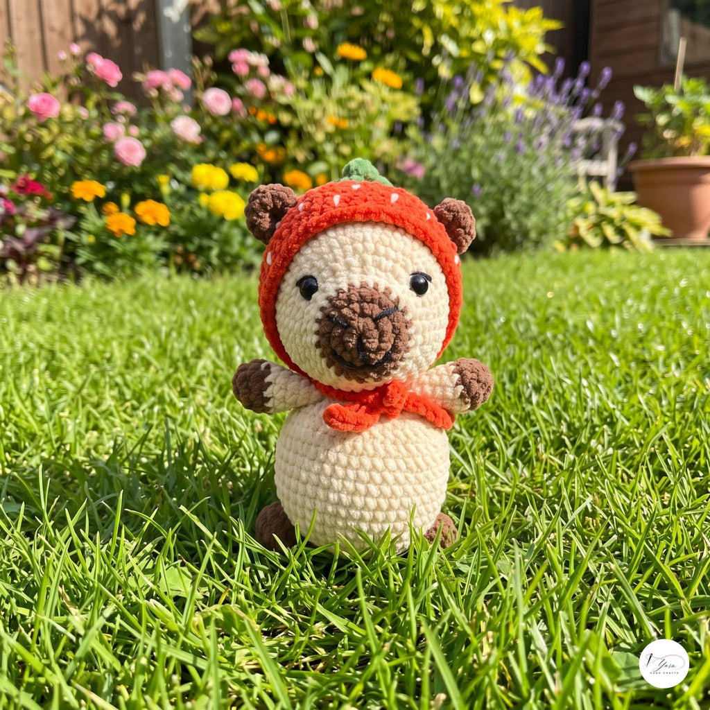 A crocheted amigurumi bear toy wearing a red winter hat and scarf, standing on a green grass lawn with a blurred garden background.
