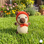 A crocheted amigurumi bear toy wearing a red winter hat and scarf, standing on a green grass lawn with a blurred garden background.