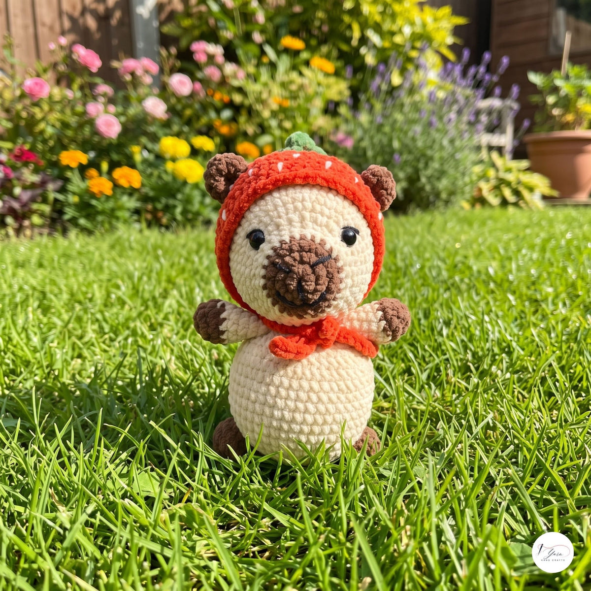 A crocheted amigurumi bear toy wearing a red winter hat and scarf, standing on a green grass lawn with a blurred garden background.