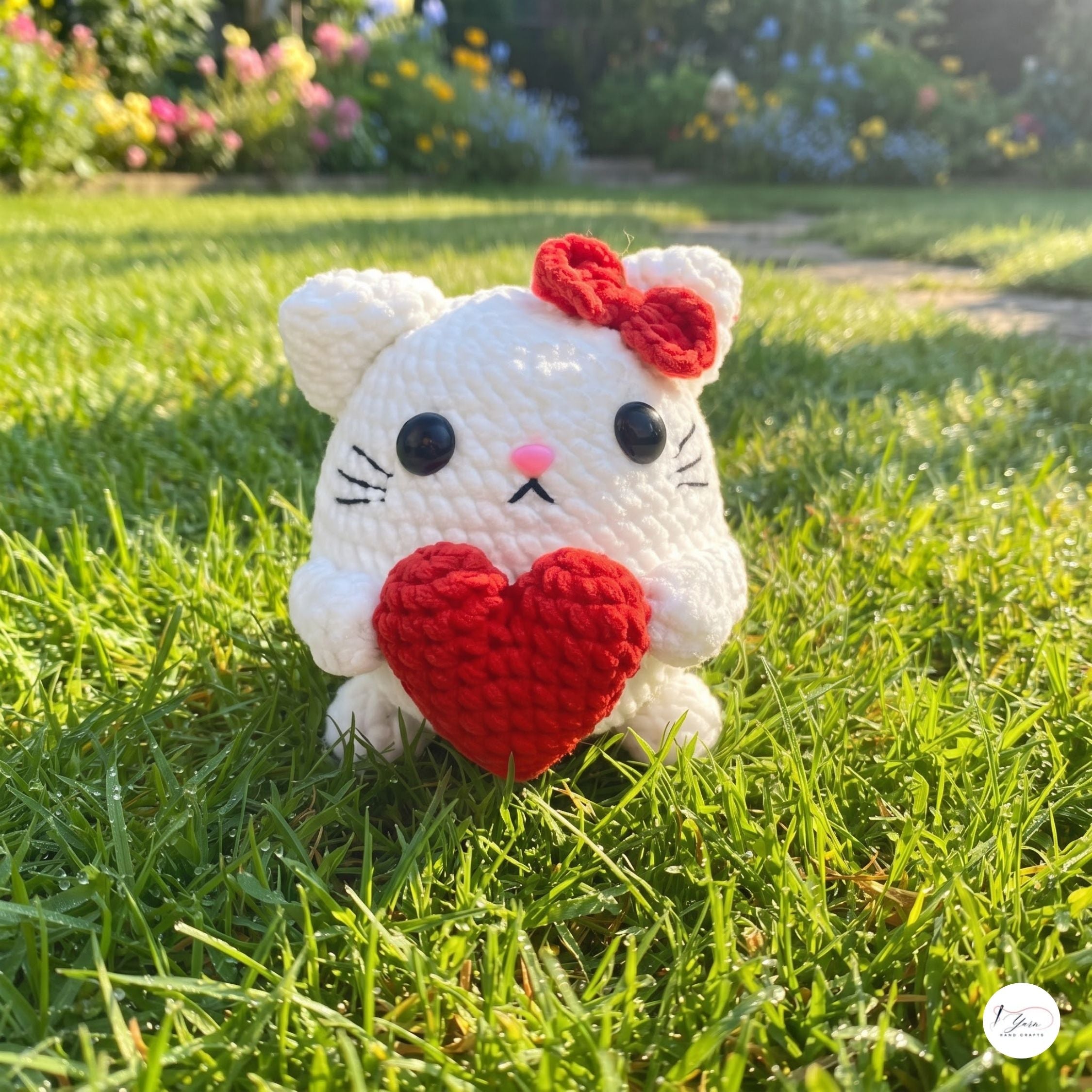 A white crocheted cat plush toy holding a red heart, sitting on a green grass lawn with a blurred garden background.