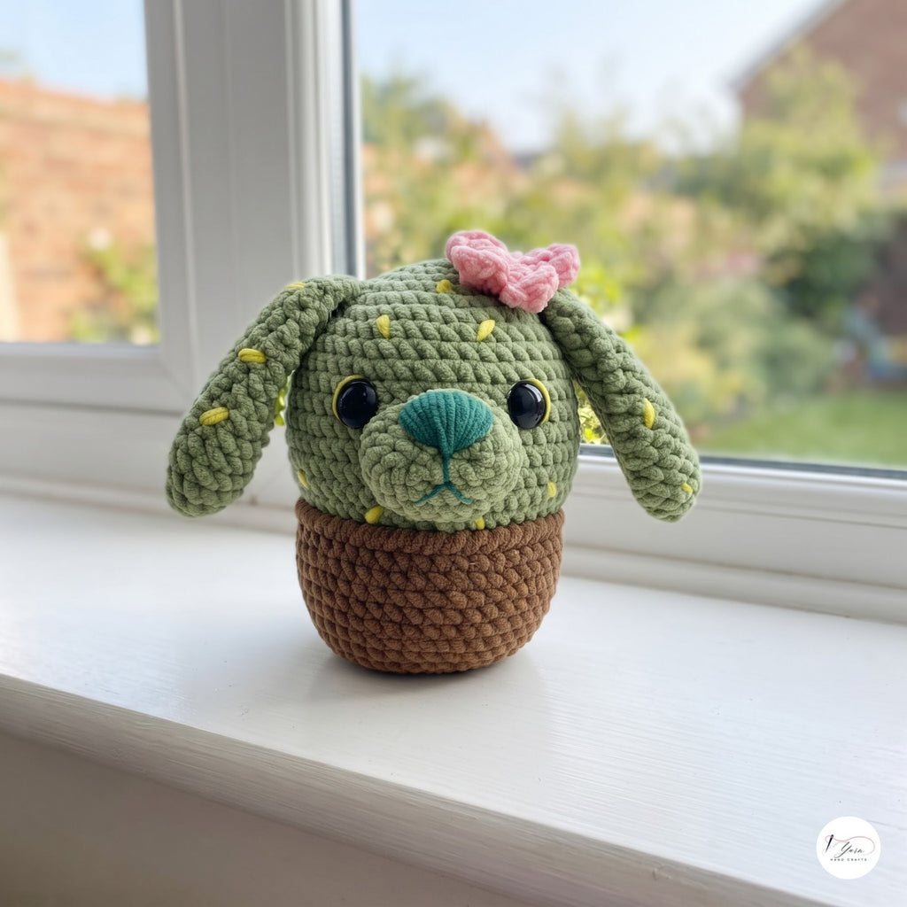A plush handmade crochet cactus puppy toy with long floppy ears, yellow embroidered spines, a pink flower, and a turquoise nose, sitting in a brown crochet pot on a white windowsill next to a window overlooking a garden.
