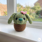 A plush handmade crochet cactus puppy toy with long floppy ears, yellow embroidered spines, a pink flower, and a turquoise nose, sitting in a brown crochet pot on a white windowsill next to a window overlooking a garden.