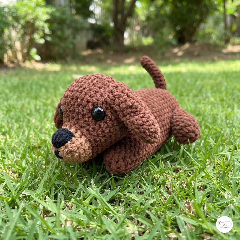 A brown crocheted plush puppy toy with black eyes and a black nose, lying on a green grass lawn with a blurred garden background.
