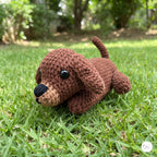 A brown crocheted plush puppy toy with black eyes and a black nose, lying on a green grass lawn with a blurred garden background.