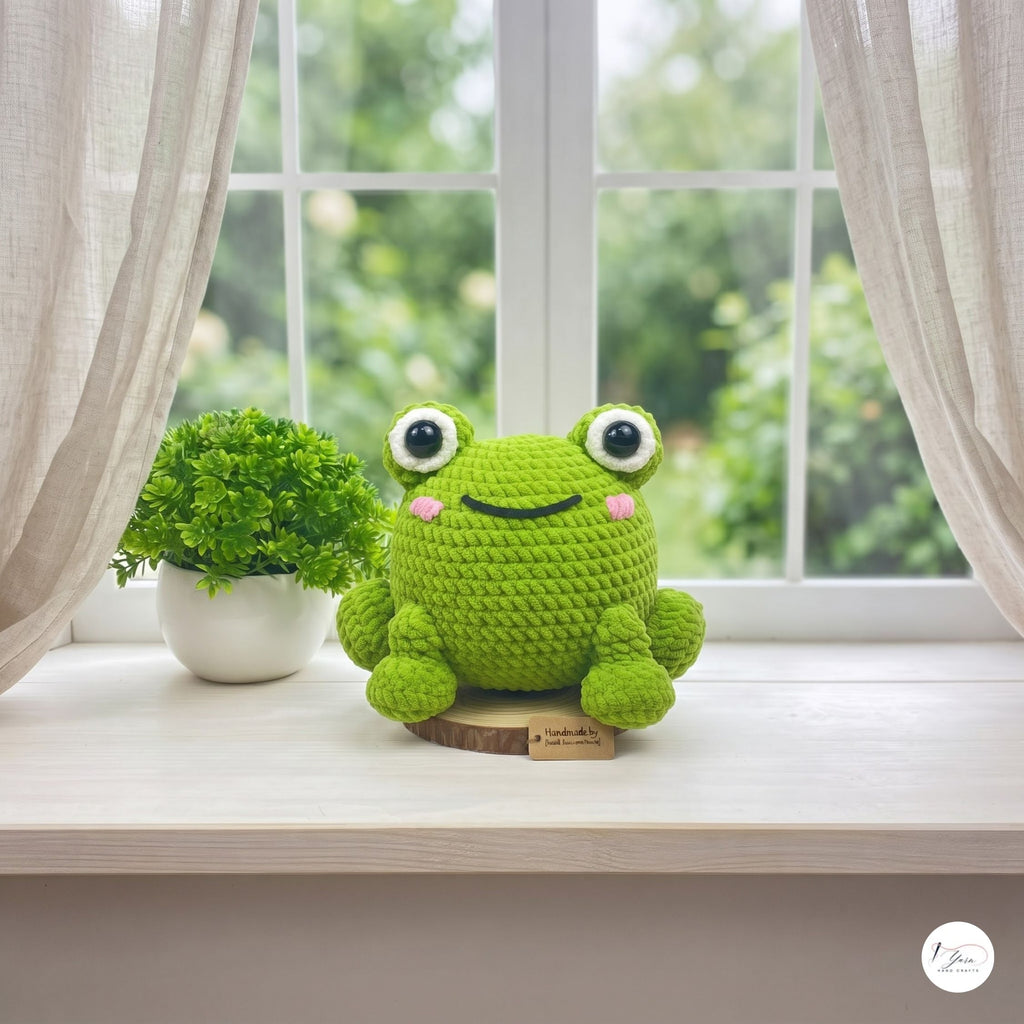 A green crocheted frog plush toy sits on a light wooden windowsill next to a small potted green plant. The window has sheer white curtains and a blurred green garden background outside.