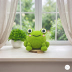 A green crocheted frog plush toy sits on a light wooden windowsill next to a small potted green plant. The window has sheer white curtains and a blurred green garden background outside.