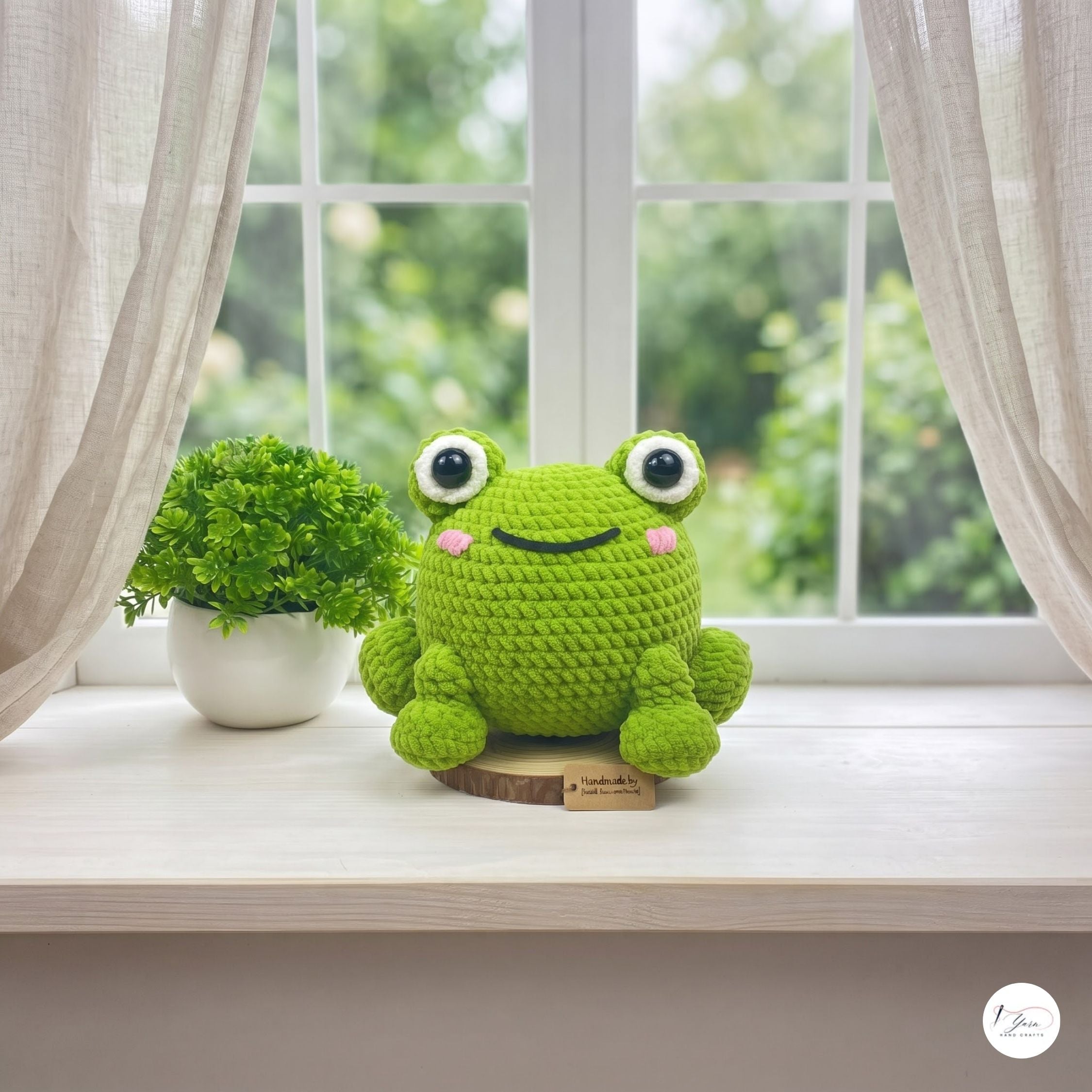 A green crocheted frog plush toy sits on a light wooden windowsill next to a small potted green plant. The window has sheer white curtains and a blurred green garden background outside.