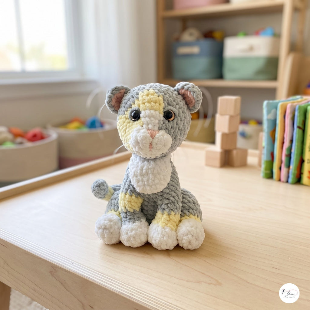 A crocheted stuffed animal toy resembling a small tiger or cat, sitting on a light wood table in a softly blurred children's room. The toy is gray with cream, white, and pale yellow accents, and has black bead eyes.