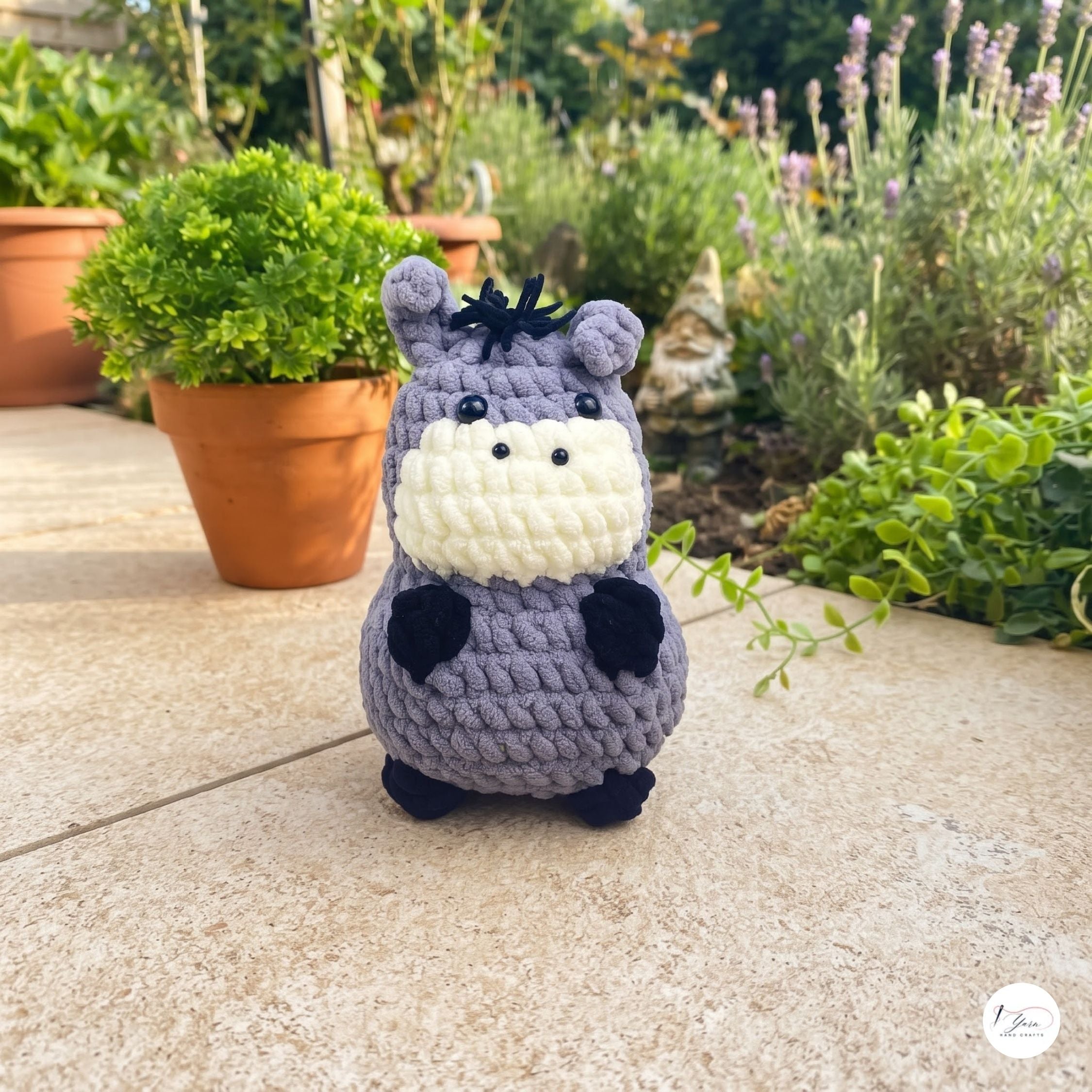 A crocheted hippo plush toy standing on a stone patio in a garden setting with potted plants and a gnome in the background.