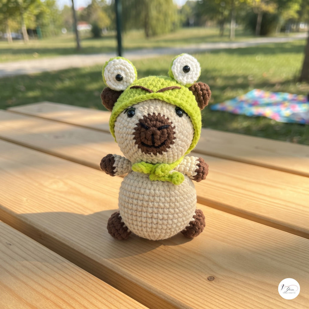 A crocheted plush toy shaped like a pug wearing a green frog hood with large white eyes, sitting on a wooden picnic table outdoors with a blurred park background.