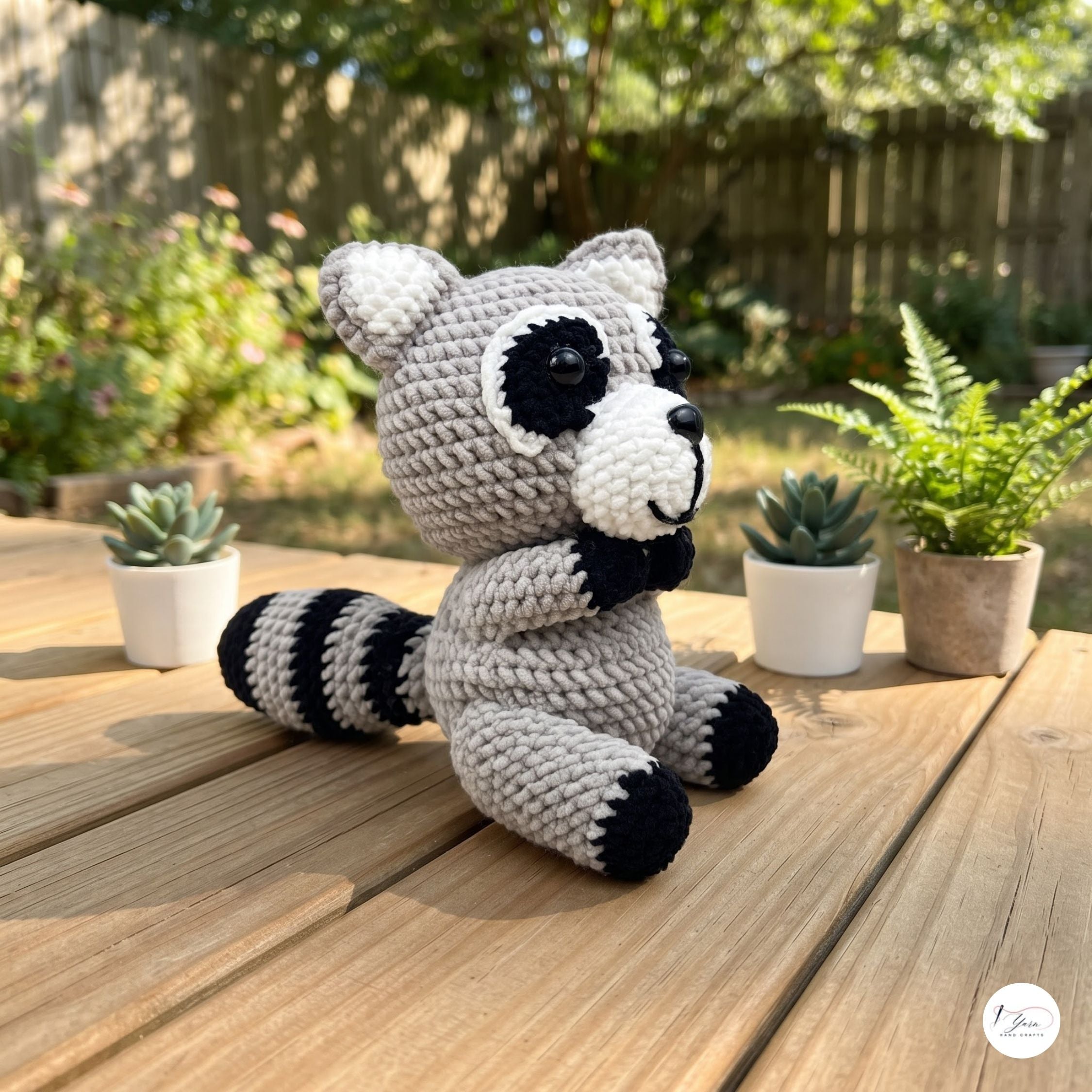 A crocheted raccoon plush toy sits on a wooden table outdoors, with small potted plants and a garden fence blurred in the background.