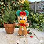 A crocheted plush character doll resembling a gingerbread figure with green ears, red bow, yellow beak, and white trim, standing on a stone patio outdoors next to a potted evergreen tree and holly plants.