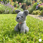 A gray crocheted dog toy sits on a green grass lawn in a sunny garden with colorful flowers in the background.