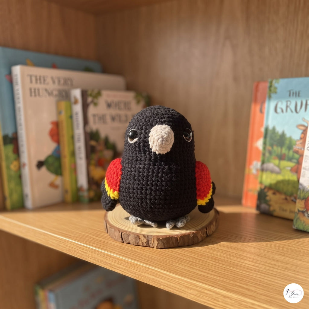 A crocheted parrot toy in black with red, yellow, and white accents, standing on a round wooden slice coaster on a wooden bookshelf with children's books in the background.