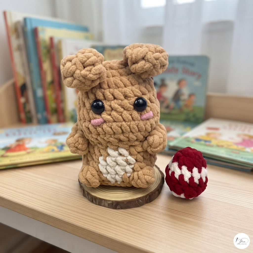 A crocheted plush bunny toy sits on a wooden tabletop in a softly lit room, with children's books blurred in the background. The bunny is tan with a white patch on its belly, black bead eyes, and small pink cheeks.
