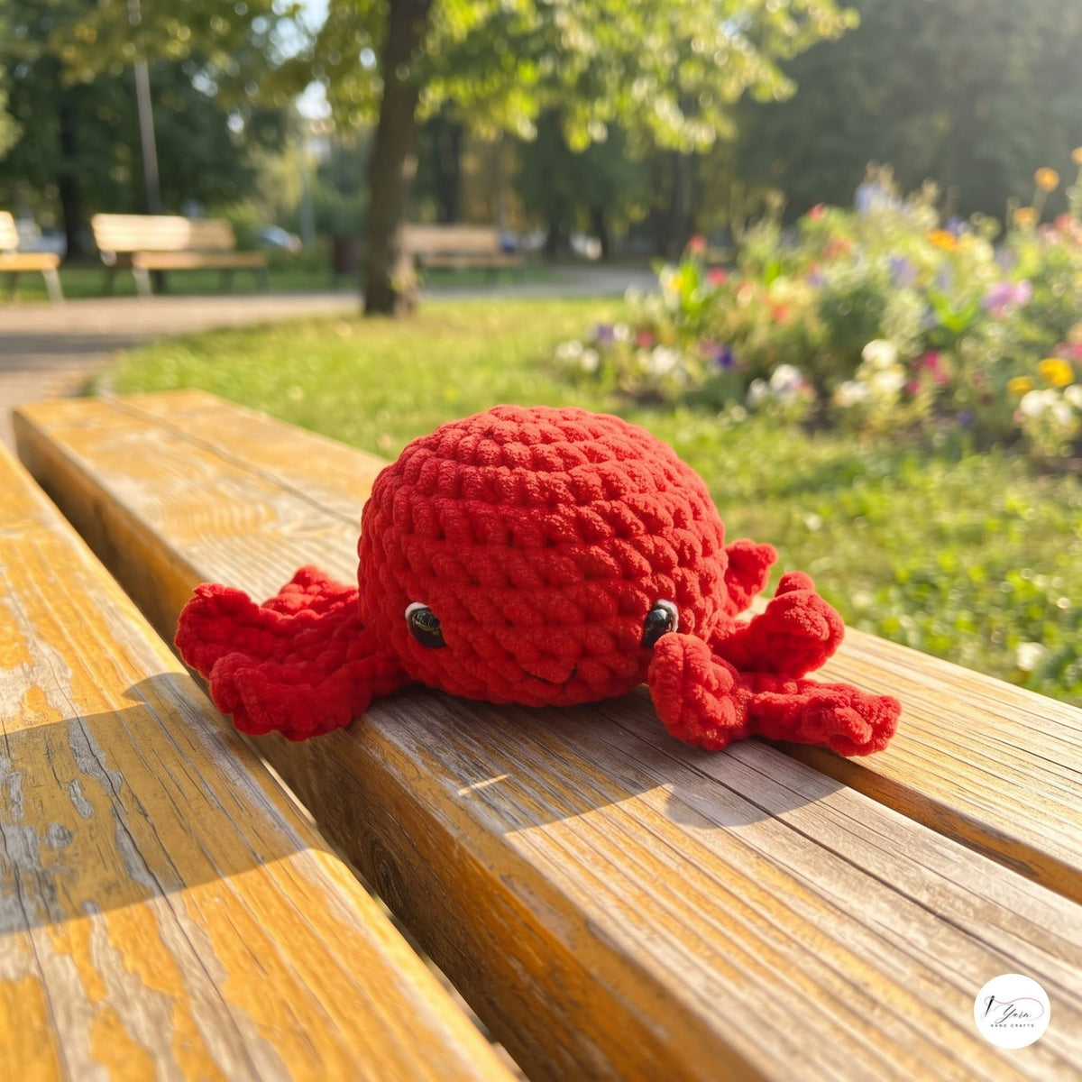 A red crocheted octopus toy with small black eyes sits on the wooden slats of a park bench, with a blurred green garden and trees in the background.
