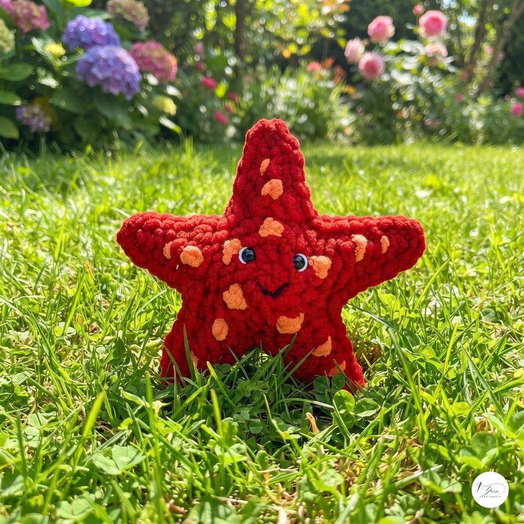 A red crocheted starfish toy with orange bumps and a smiling face, placed on a green grassy lawn with a blurred garden background.