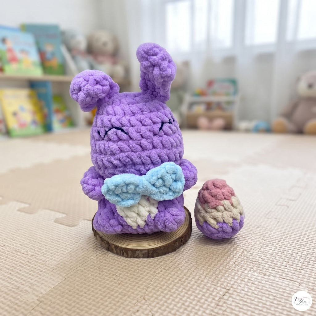A crocheted purple bunny figurine with a light blue bow and white wing details, standing on a small wooden slice next to a matching crocheted egg, set on a beige play mat in a softly blurred room.